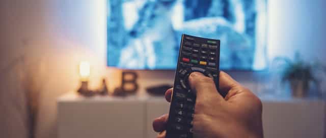 Eine Hand hält eine Fernbedienung. Man sieht im Hintergrund einen Fernseher.